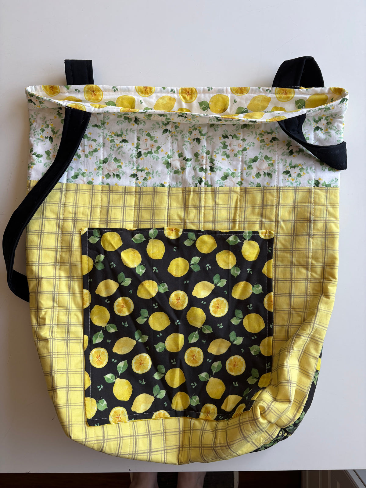 A handmade quilted tote bag featuring a yellow and white lemon print, a white floral patterned section, and a yellow plaid border, with black shoulder straps.